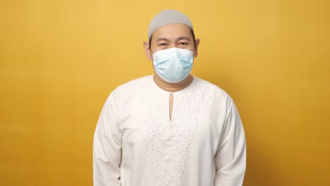 portrait of asian muslim man wearing facial mask, against yellow background, new normal due to covid coronavirus pandemic