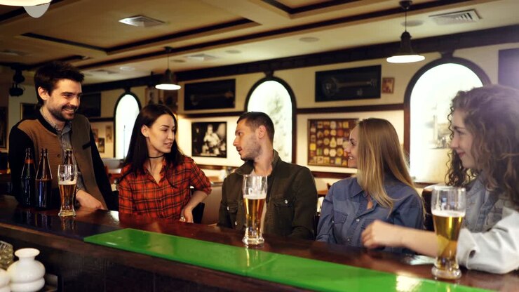 Un gruppo di amici è seduto al bancone di un bar e parla. Bicchieri e bottiglie di birra, tavoli sono visibili. Atmosfera amichevole e informale