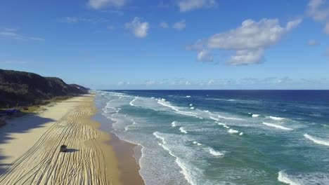 beautiful cinematic descending shot over the beach of fraser island as a solitary four-wheel drive car drives up the beach among the many tracks left by other vehicles