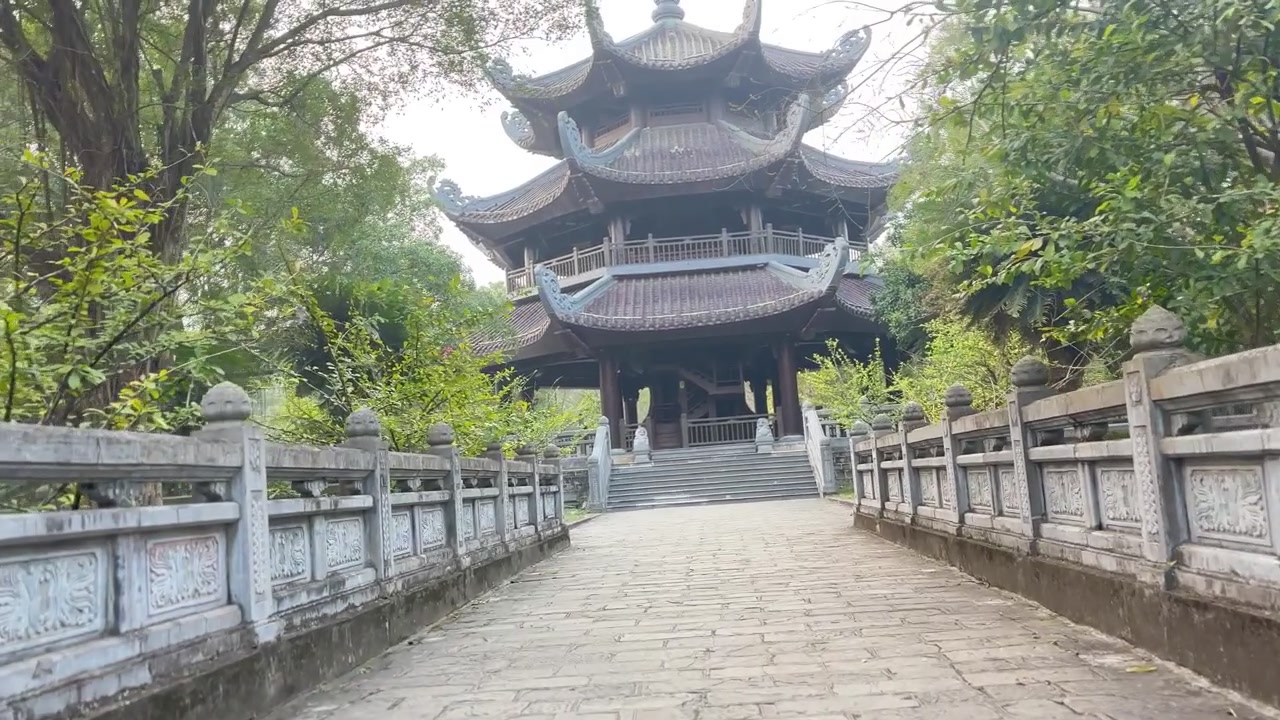 Naturskøn udsigt over traditionel fleretagers træpavillon ved Bai Dinh Pagoda i Ninh Binh, Vietnam, omgivet af frodige grønne træer og et fredfyldt tempelstiområde.