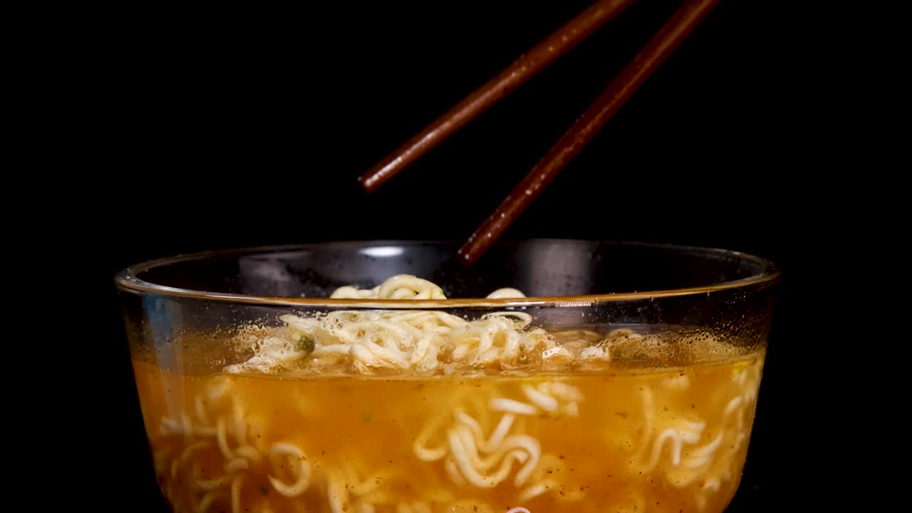A hand uses chopsticks to stir and lift cooked instant noodles in a glass bowl filled with broth, against a black background with studio lighting