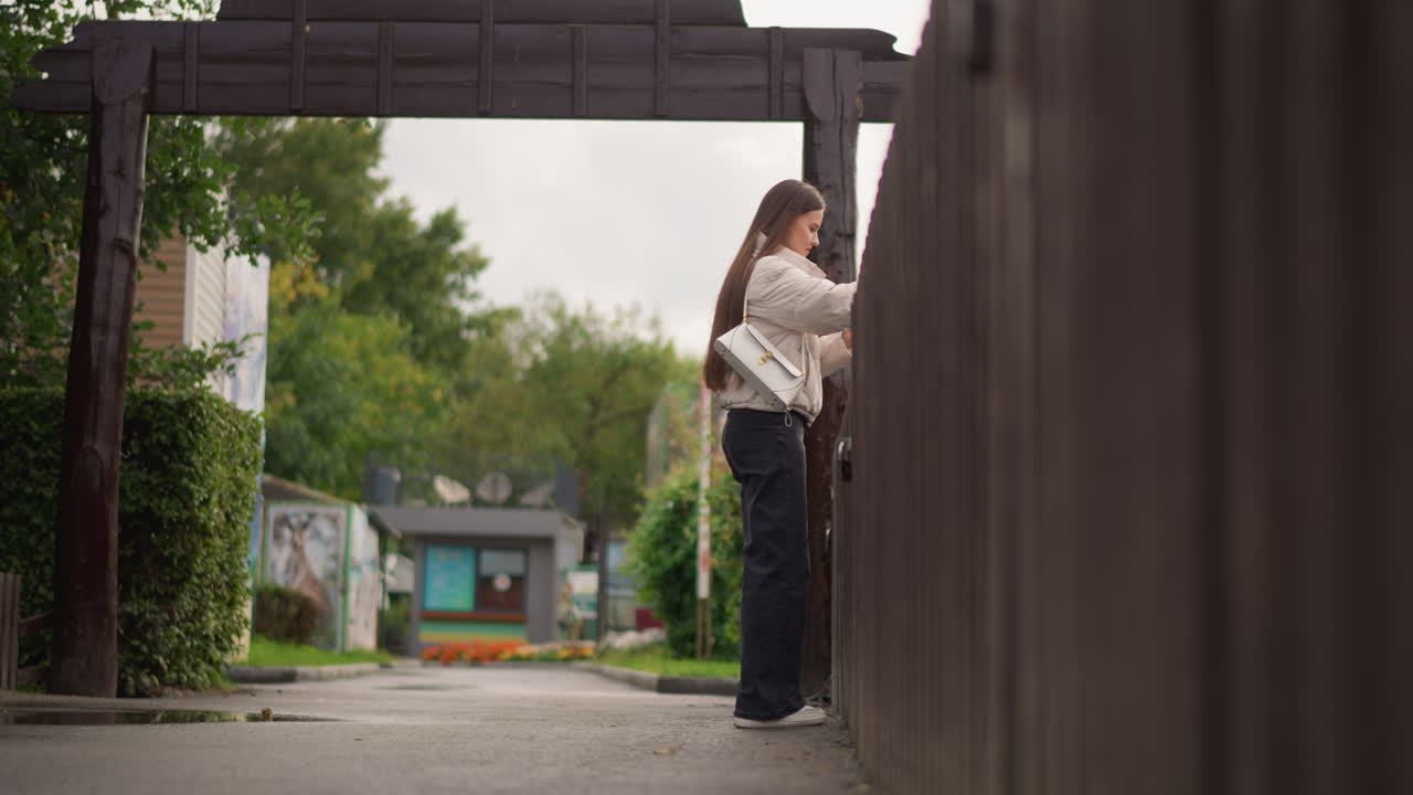 young woman standing at wooden gate reaching to open latch, framed by suburban alley with mural on background building, overcast sky, casual outfit with jeans and jacket, sneakers, soft natural