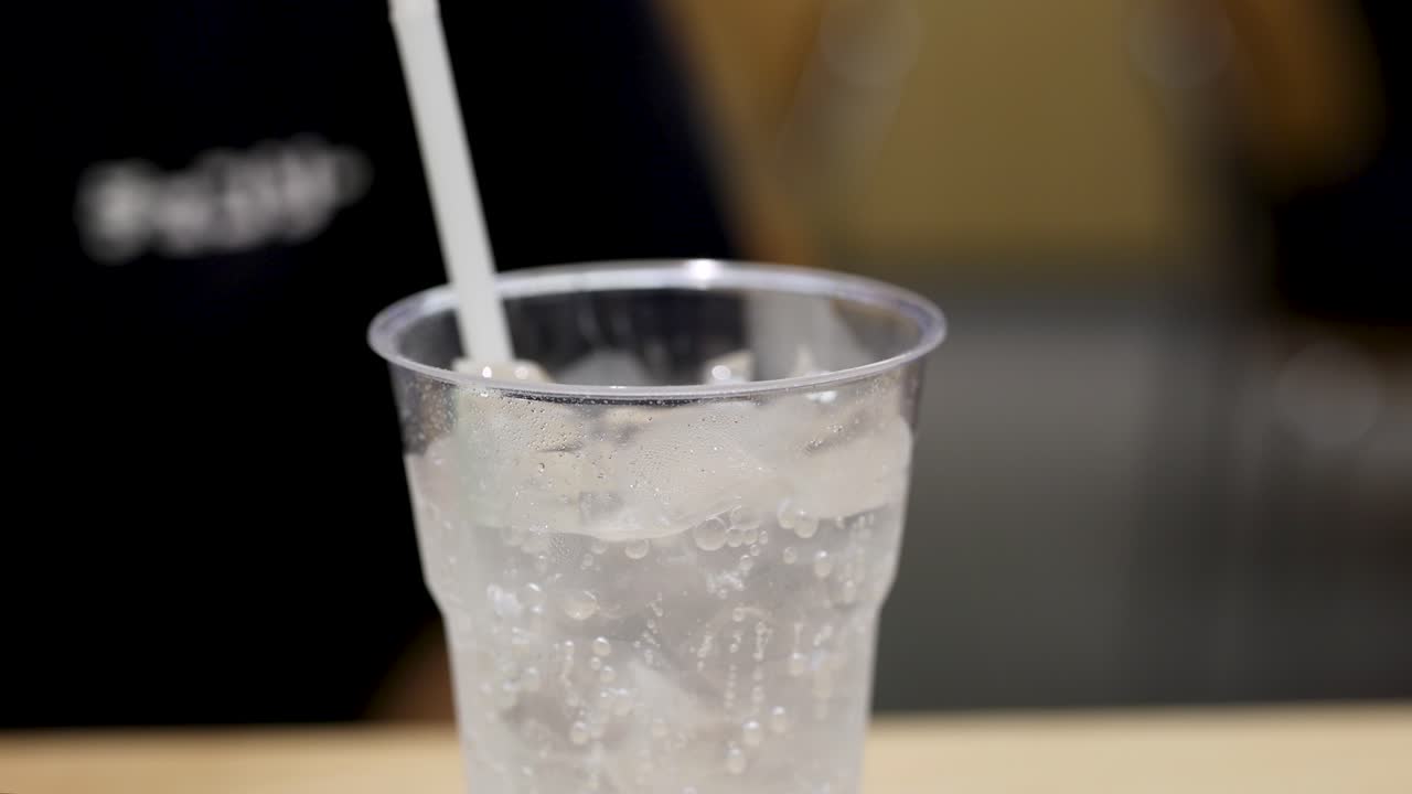 Plastic cup of cold carbonated drink with ice and straw, indoor setting, soft lighting