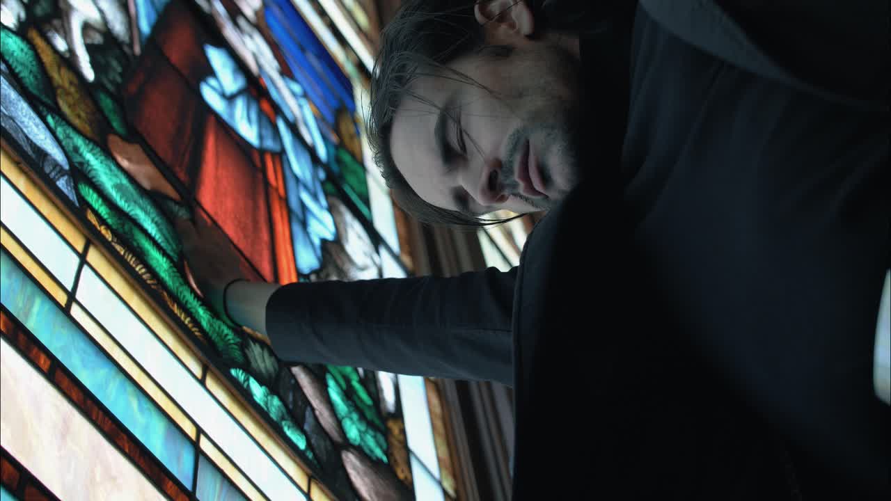 Close-up of a man reaching out to touch a stained-glass church window. The shot captures intimacy, faith, and reflection, highlighting the vivid colors and sacred atmosphere