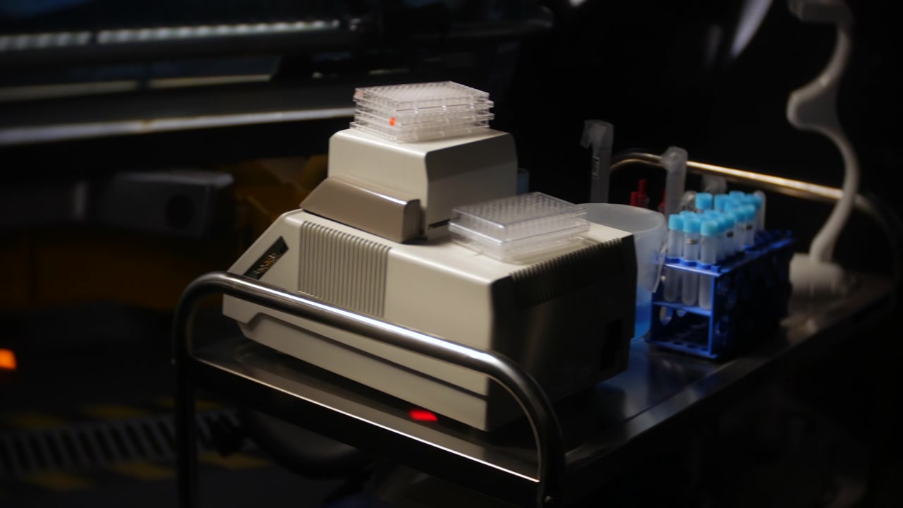 Laboratory analyzer beside test tubes and sample trays on a stainless steel cart. Dark biotech workspace with medical research equipment and diagnostic tools