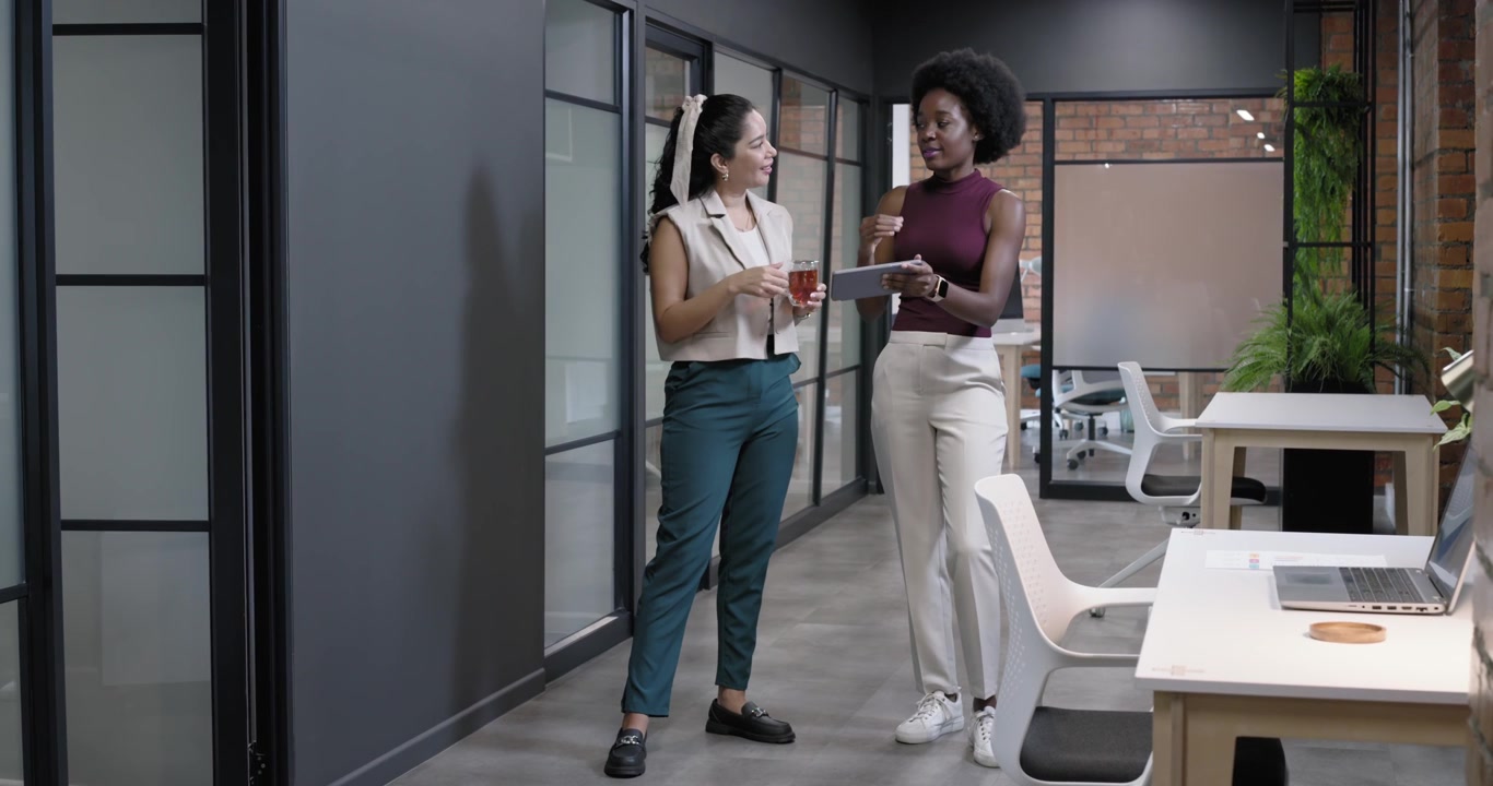 Female coworkers standing at office right woman tapping tablet left woman with glass reviewing work