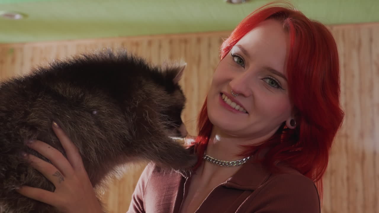 Zrzavá žena s bílou pletí objímající mývala v útulném pokoji obloženém dřevem, něžné objetí, usměvavý výraz, srst přitisknutá k rameni, moment záchrany domácího mazlíčka s jemným teplým osvětlením a intimním portrétem
