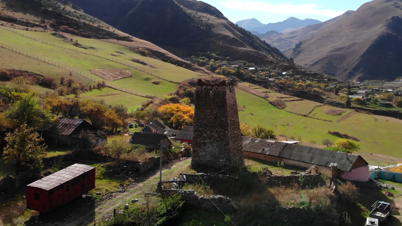 Ampia vista aerea di una rovina di un'antica torre in pietra che domina una valle montana rurale con campi verdi, case sparse e colline distanti in piena luce diurna