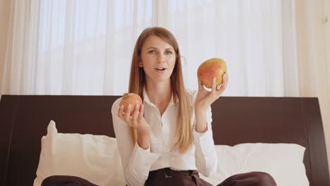 pleasant caucasian woman holding pomegranate and mango in hands and talking on camera. travel blogger sitting on comfy bed at bright hotel room. woman holding pomegranate and mango