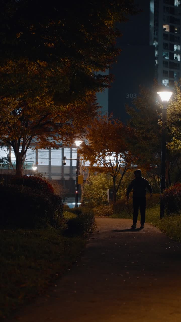 Vertical shot of a silhouetted man in a hoodie walking along a park path at night after exercising, surrounded by autumn trees illuminated by streetlights
