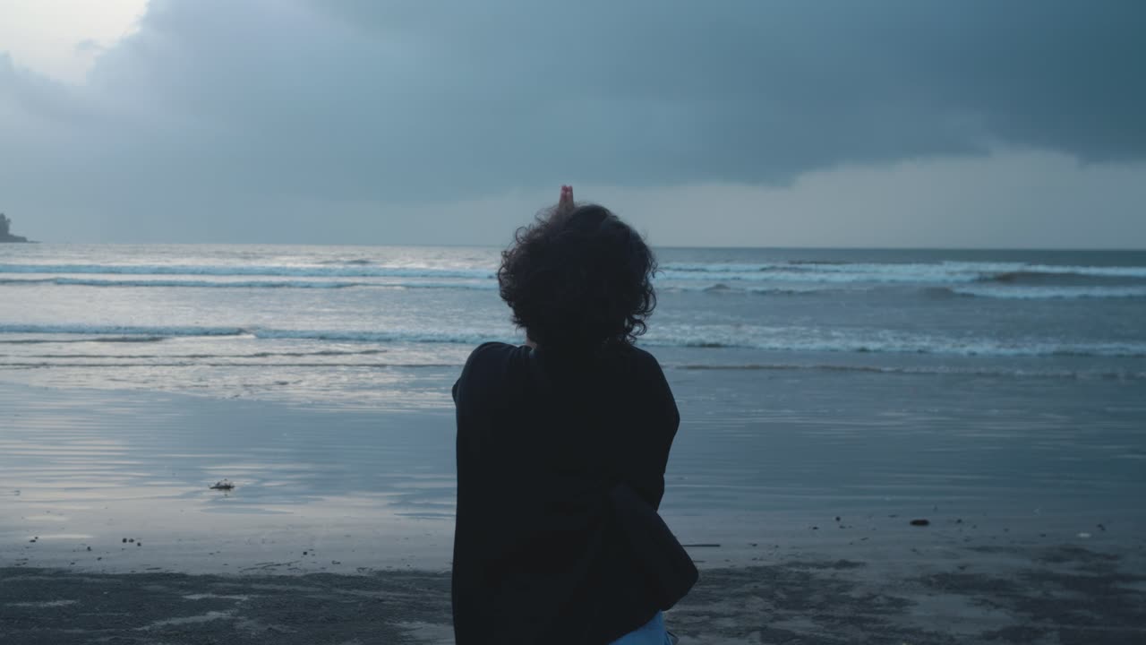 Back view of woman on sandy beach praying with folded hands as small waves move gently in the background