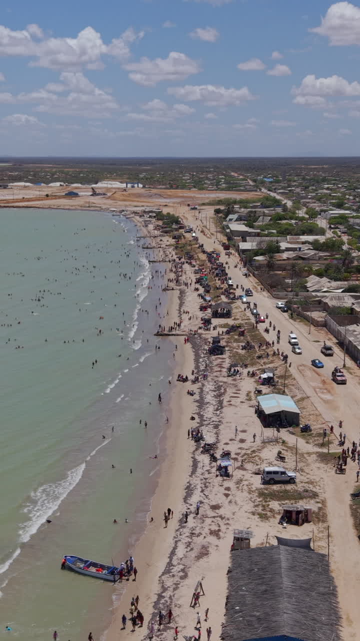 Video dọc trên không bằng drone ghi lại bãi biển sôi động của Manaure ở La Guajira, Colombia với đám đông, hoạt động sôi nổi trên bờ biển, nước màu ngọc lam, những túp lều mộc mạc và ánh nắng nhiệt đới.