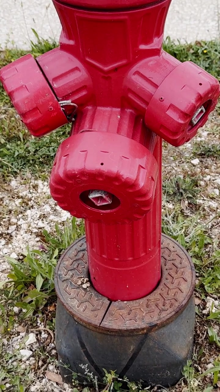 Primo piano di un idrante antincendio rosso all'aperto, che mostra attrezzature per l'accesso all'acqua di emergenza accanto a erba e pavimentazione in un'area pubblica