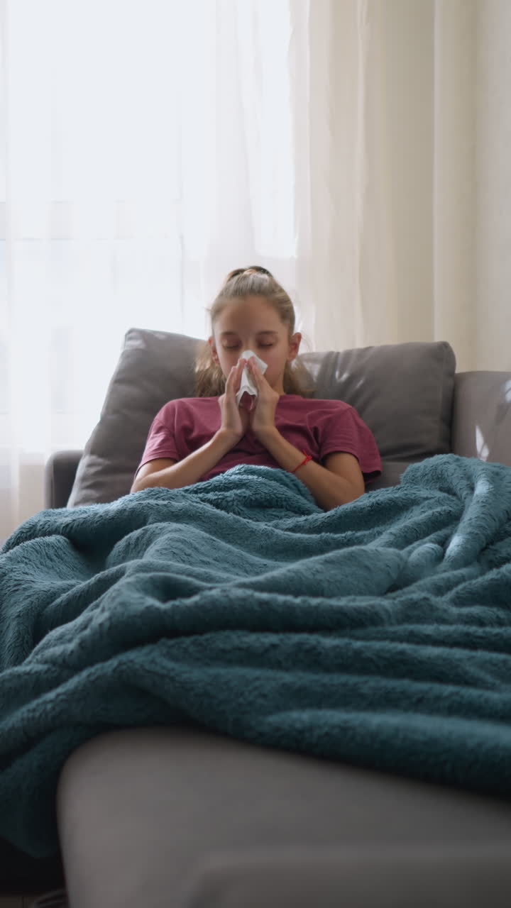 Ragazza in rosa con l'influenza sdraiata sul divano, che starnutisce nel fazzoletto con medicinali e un bicchiere sul tavolo, avvolta in una coperta, con un aspetto esausto e malato