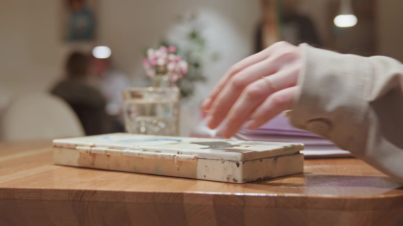 Nahaufnahme einer Hand, die einen alten Malkasten auf einem Holztisch bei sanfter Beleuchtung platziert. Ein lila Notizbuch und ein Wasserglas stehen daneben, mit verschwommenen Gästen, die an Tischen hinter dem Hauptmotiv sichtbar sind. Die Szene zeigt
