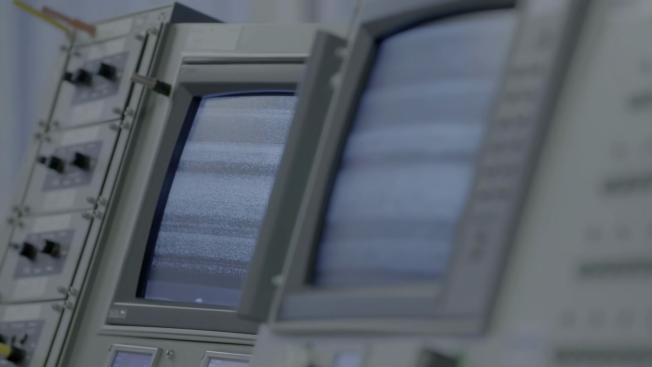 Close angled view moves from sharper foreground screen toward second monitor, revealing flickering gray noise, horizontal bands, pale equipment housings, buttons, and control knobs.