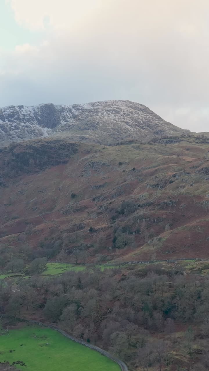 Vertical forward drone flight toward a distant mountain with a small snowy peak. Green and brown terrain rises in the beneath large white clouds and a slightly gloomy sky, creating a moody atmosphere