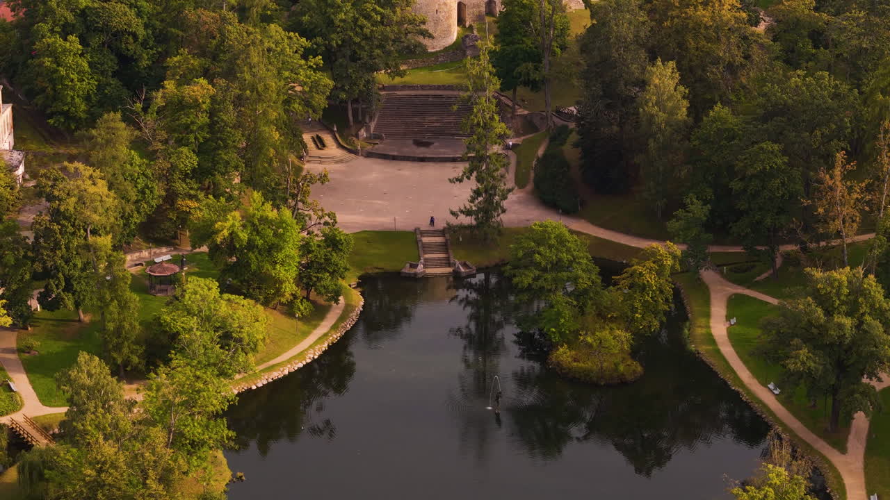 Aerial footage of Cesis Medieval Castle in Latvia, surrounded by lush greenery and a scenic pond.