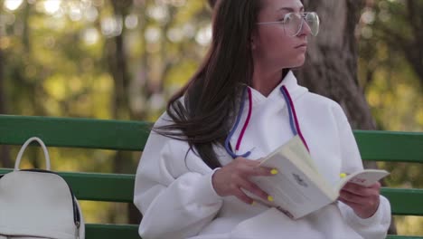 Chica-Con-Gafas-Leyendo-Un-Libro-En-Un-Banco-En-Un-Ambiente-Tranquilo-En-Un-Parque