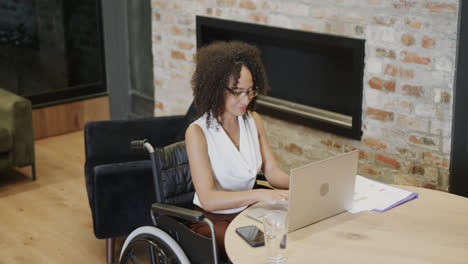 feliz mujer de negocios biracial en silla de ruedas usando portátil en el escritorio en la oficina, en cámara lenta