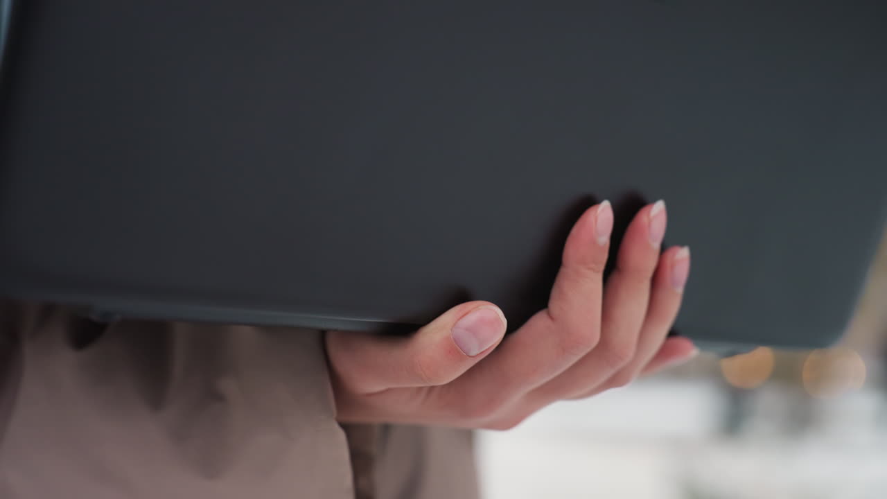 Close-up of light-skinned hand gently holding black laptop while fingers subtly move, soft winter background with blurred lights creating calm atmosphere of outdoor focus and technological engagement