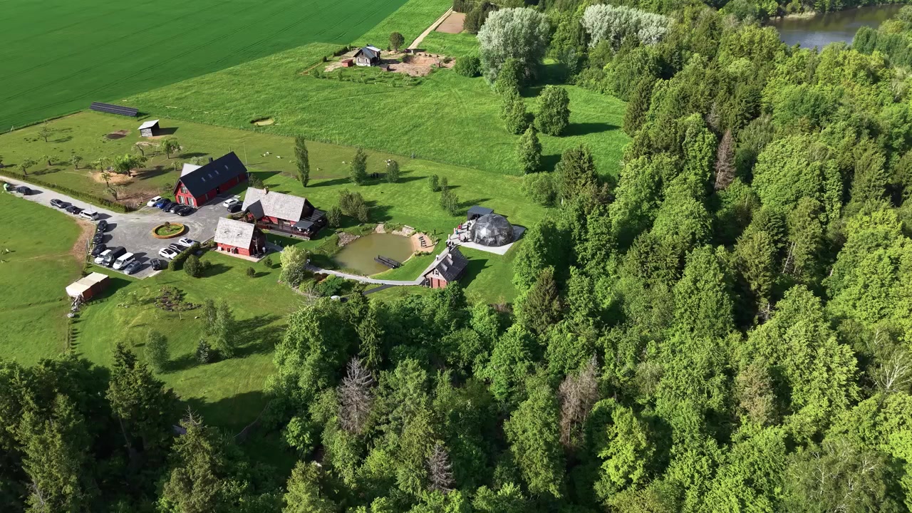 Elokuvallinen dronevideo tallentaa rauhallisen maaseututilan punaisine maalaistaloineen, ympäröivine puineen, lammikoineen ja rehevine vihreine peltoineen kirkkaassa päivänvalossa. Tasainen ilmakuvaus näyttää elävää, luonnonkaunista maisemaa.