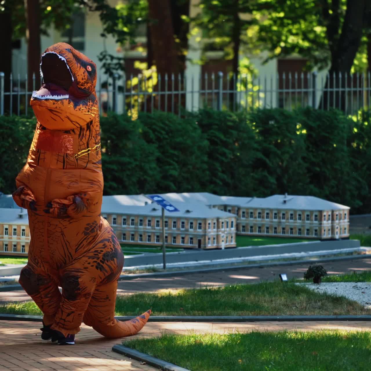 Cartoon animation. Dragon costume in small city. Person wearing blow-up animal suit walking among beautiful handmade architecture in the park.