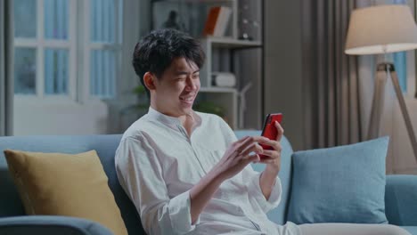 Asian Man Touching Smartphone And Laughing While Sitting On Sofa In The Living Room
