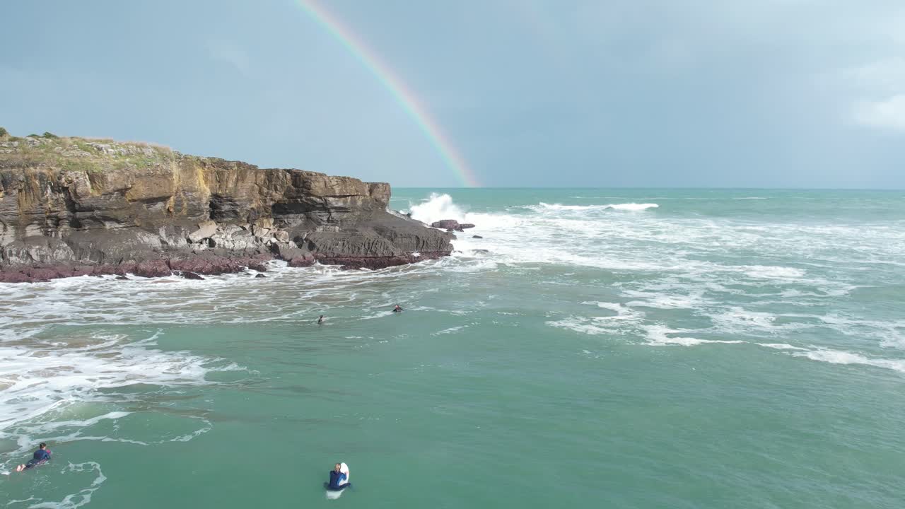 gente irreconocible mirando las olas rompiendo en los acantilados, con un arco iris en el fondo