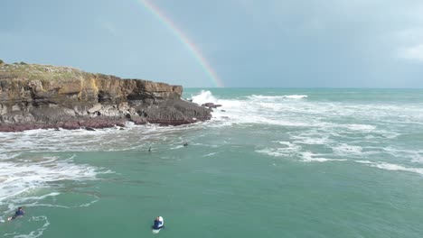 Gente-Irreconocible-Mirando-Las-Olas-Rompiendo-En-Los-Acantilados,-Con-Un-Arco-Iris-En-El-Fondo