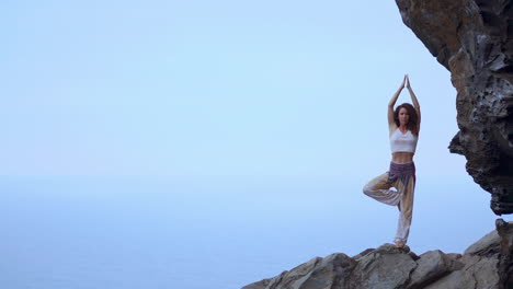 At-sunset-by-the-blue-ocean,-a-young-woman-engages-in-yoga-on-a-rocky-seashore,-portraying-the-concept-of-a-healthy-lifestyle,-harmony,-and-the-interconnectedness-of-humans-and-nature