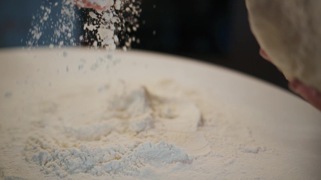 Una mano sparge farina sul tavolo mentre tiene l'impasto. La calda luce della cucina illumina la polvere di farina. Viene mostrato il processo di preparazione del pane o della pasta. Ideale per progetti di cucina, pasticceria o legati al cibo.