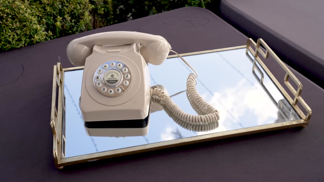 Sebuah telepon vintage terletak di atas nampan reflektif. Latar menunjukkan langit cerah dan beberapa tanaman hijau di latar belakang, bersama dengan perabot di dekatnya