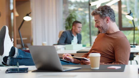 casually dressed mature businessman with feet on desk in office using mobile phone making notes