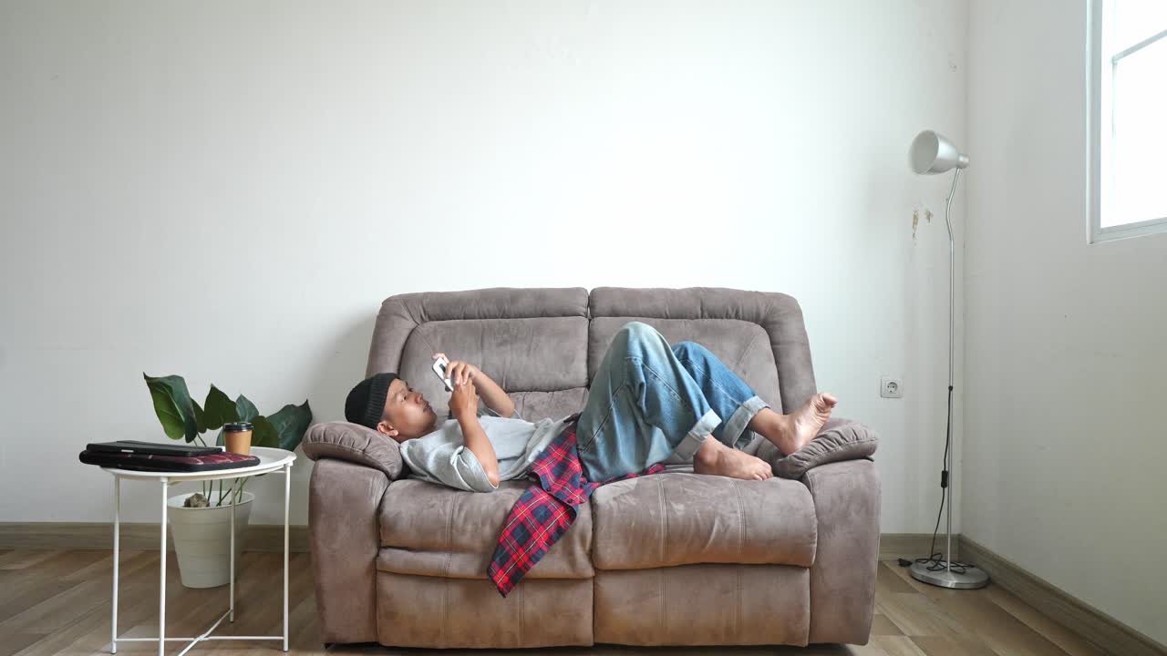 Young Asian College Student In Casual Style Using A Smart Phone Lying On Couch At Home