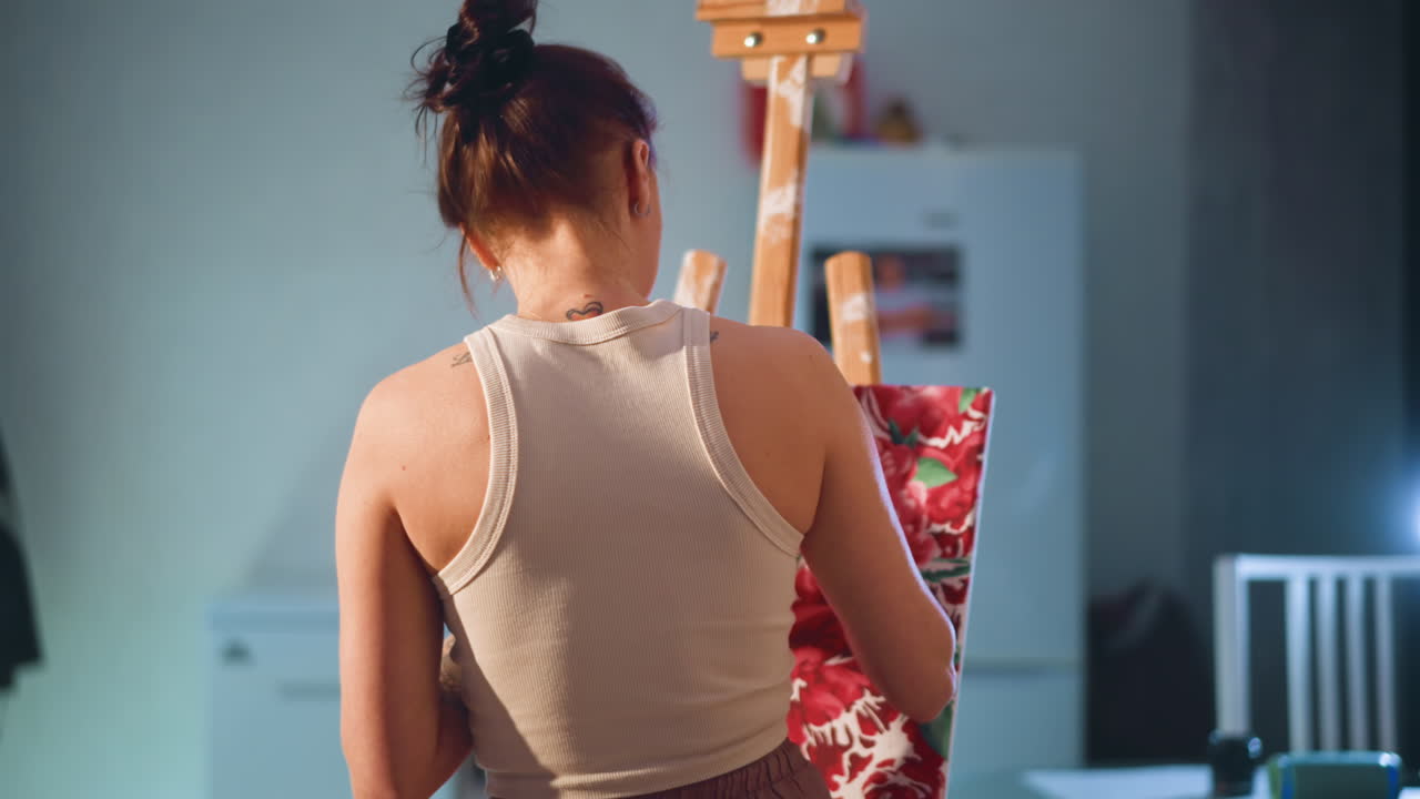 Back View Caucasian Woman Painting Easel In Cozy Home Studio, Sleeveless Top, Hair Bun, Soft Warm Light, Refrigerator And Chair In Background, Focused Strokes On Canvas, Quiet Creative Atmosphere