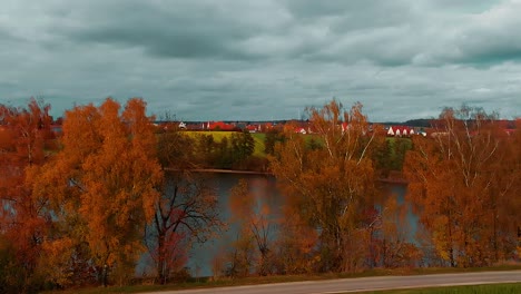 Un-Panorama-Cielo-Nublado-Techos-Rojos-De-Un-Pequeño-Pueblo-Prado-Y-Hermosos-árboles-Con-Colores-Otoñales-A-Lo-Largo-Del-Lago