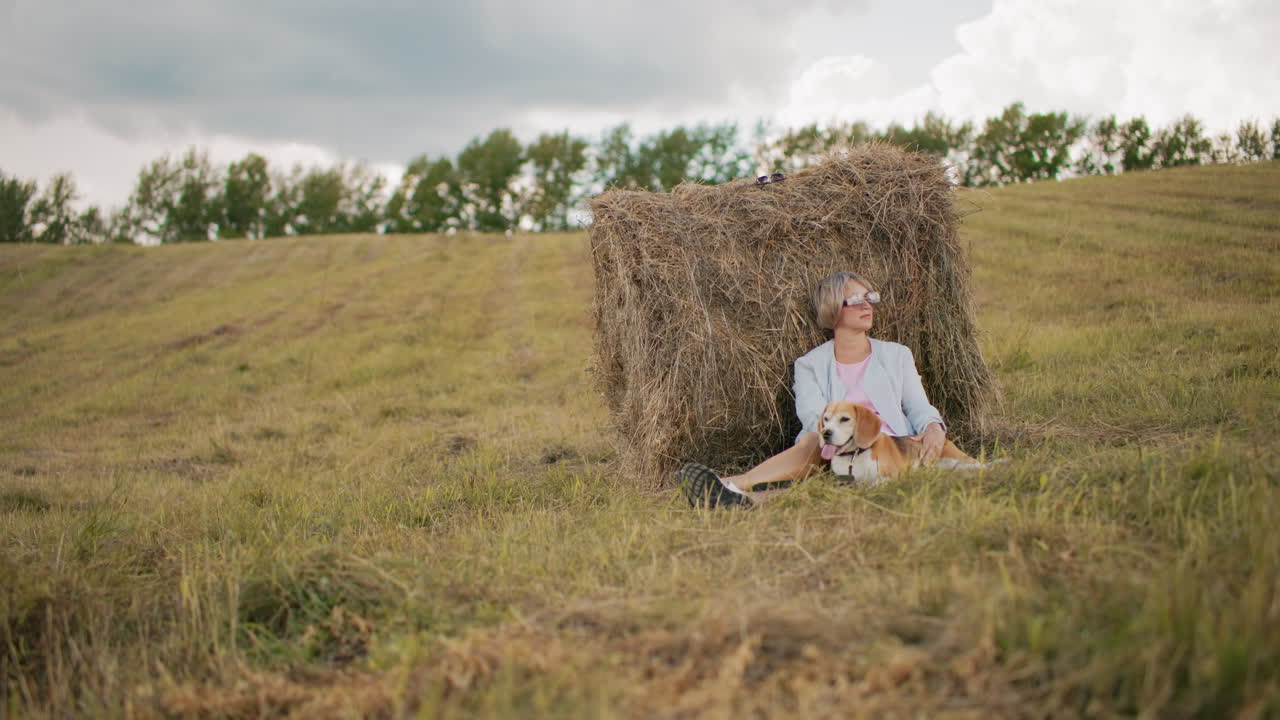 женщина в солнцезащитных очках и повседневной одежде сидит на травяном поле, опираясь на сено, мягко лаская собаку, открытая сельская местность с холмистыми холмами, облачным небом и мирной сельской обстановкой