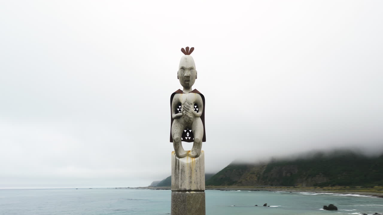 흐린 날 뉴질랜드의 큰 만 앞에 있는 토템 기둥 꼭대기에 있는 전통 마오리 동상의 공중 팬 샷