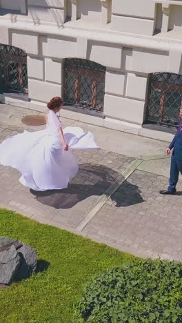 happy girl in long wedding dress dances on street and groom watches near elegant buildings on sunny day aerial view