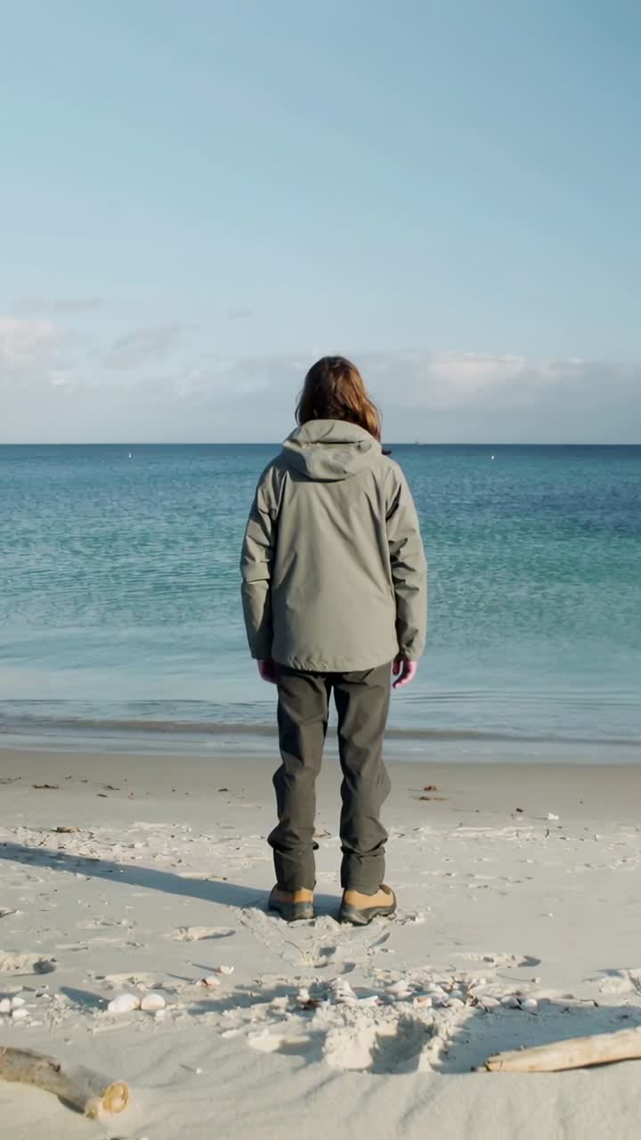 Vertical video: Standing adult in hooded jacket shifting weight as waves lap sand at beach, boots
