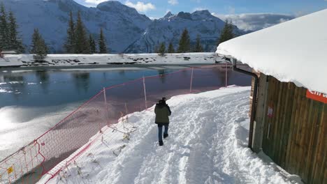 Walking-between-a-lake-and-log-cabin,-on-a-snow-laden-pathway,-located-on-a-mountain-winter-getaway-at-Engelberg,-Brunni-in-Bahnen,-Switzerland