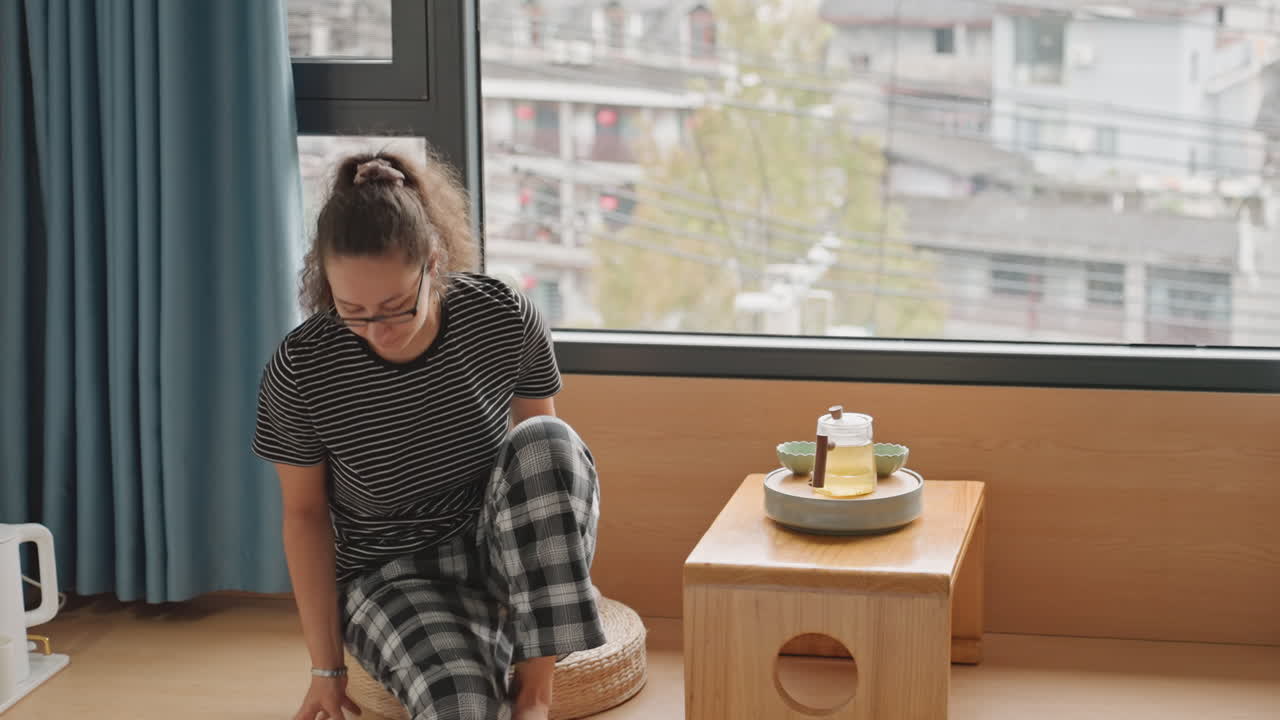 Ospite femminile che si gode tè e torta in hotel su una panca vicino alla finestra, sistema tazza e piatto con un sorriso gentile sullo sfondo di una vista urbana. Arredamento minimalista con tavolo in legno e cuscini, atmosfera di relax da 'staycation'.