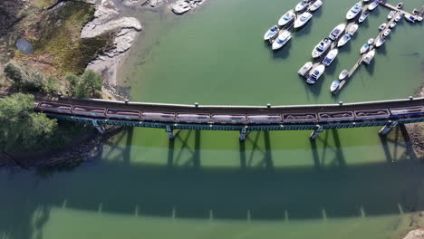 Aerial-birds-eye-view-of-freight-train-pulling-cargo-carridges-across-bridge-over-water-UK-4K