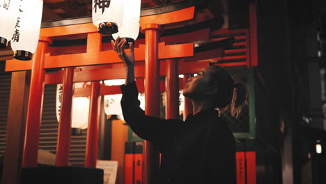 Woman-dressed-in-black-reaches-up-to-touch-an-illuminated-lantern-in-a-Japanese-shrine