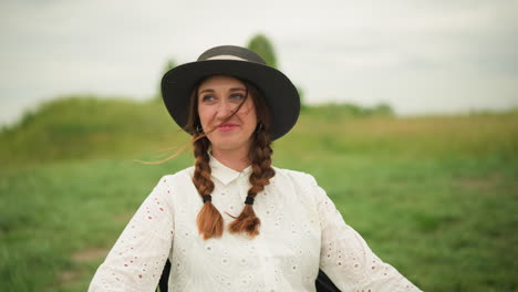 a woman wearing a white dress and a black hat is sitting in a green field, smiling warmly. the outdoor setting highlights her joyful expression, a moment of happiness in a natural environment