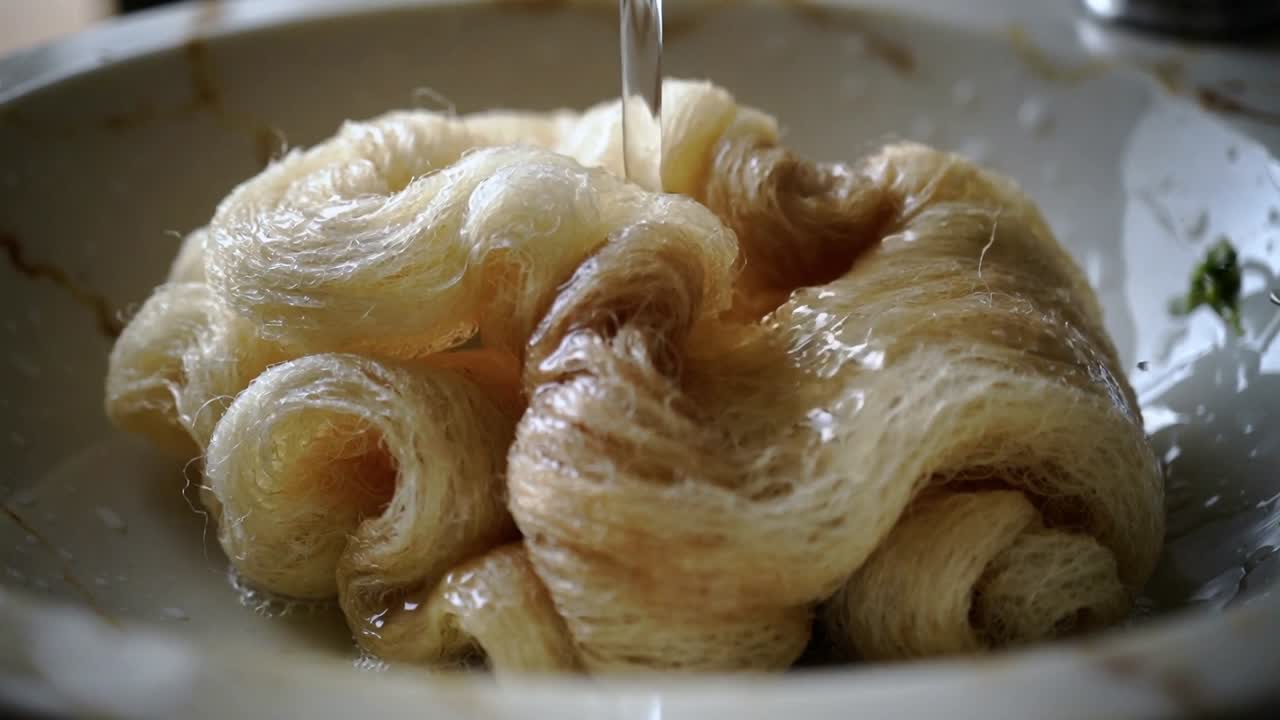 Un getto d'acqua bagna fili arrotolati in una ciotola con bordo decorato su un bancone da cucina, formando una pozza.