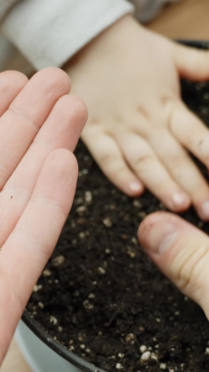 Close-up vertical frame showing a child and adult working together to plant in soil. Their hands press into the dirt, symbolizing care, learning, growth, and human connection through nature.