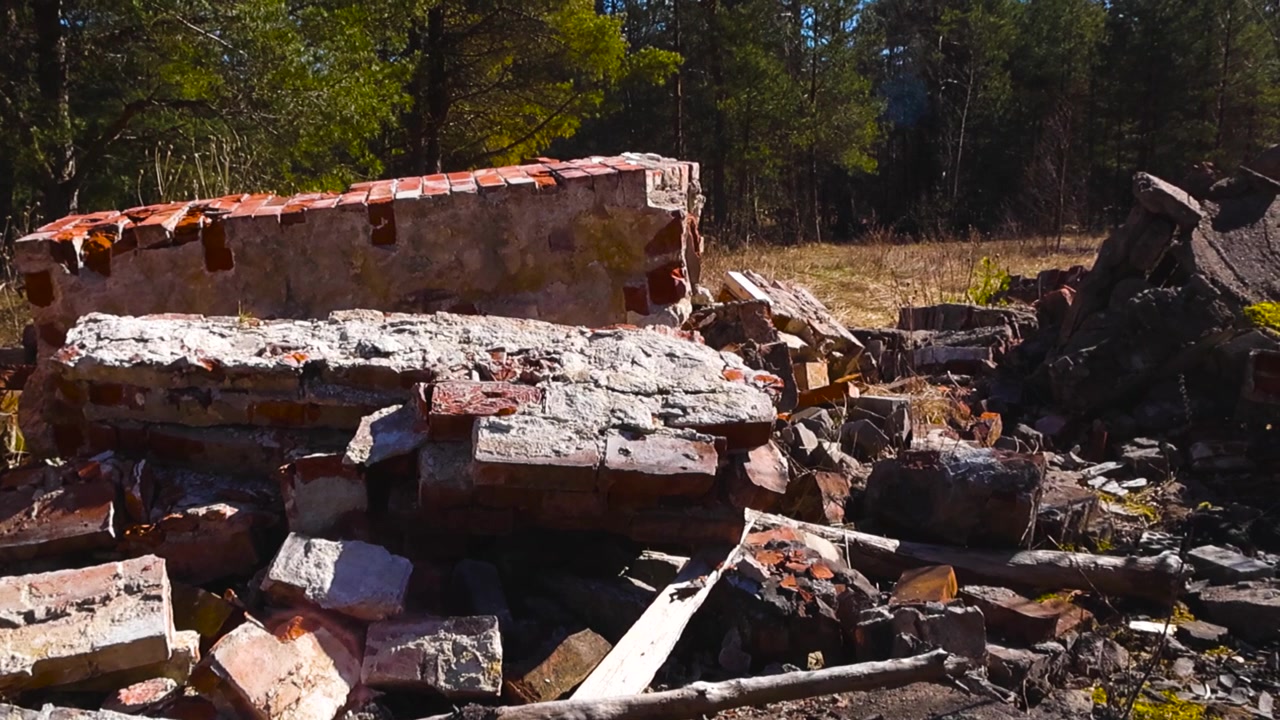 Raggi di sole e ombre che si muovono attraverso le rovine di un edificio in mattoni rossi crollato, rivelando un mucchio di detriti stagionati abbandonati in una radura tranquilla circondata da una fitta pineta.