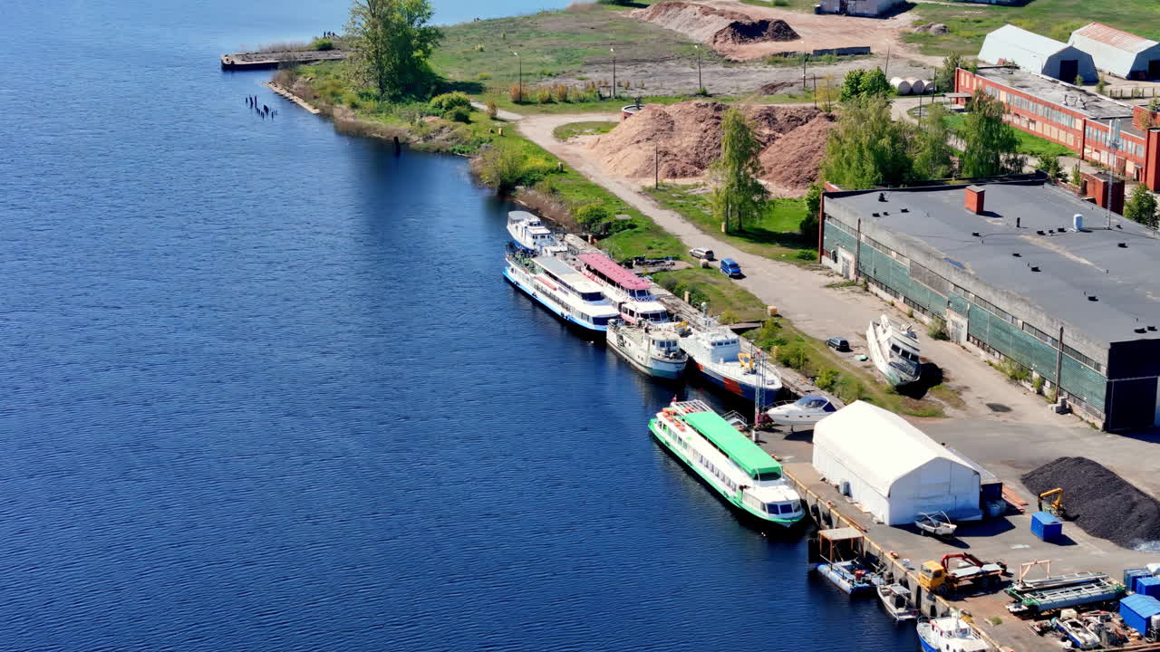 Elevated Aerial View of a Riverside Industrial Area with Various Moored Boats, Including Passenger Vessels and Yachts, Alongside Warehouses, Piles and a Distant Grassy Shoreline on a Bright Sunny Day